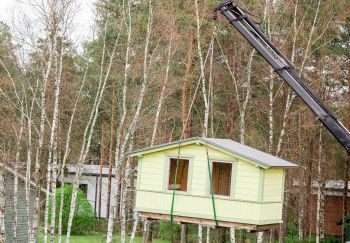 Small house being lifted by a crane in a wooded area with bare trees